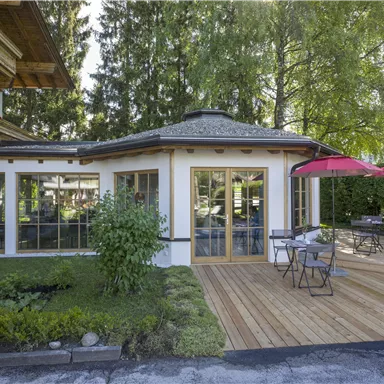 Eine gepflegte Terrasse mit Holzbelag und Sicht auf einen gemütlichen Garten. Im Hintergrund steht ein Haus mit großen Fenstern und einem markanten Dach.