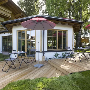 Ein gemütlicher Außenbereich mit Holzterrasse und Tischen. Zwei rote Sonnenschirme spenden Schatten in einer schönen Gartenlandschaft.