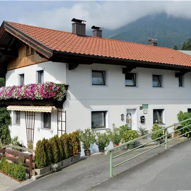A charming house with a red tiled roof and colorful flowers on the balcony. In the background, gentle hills and a misty mountain landscape can be seen.