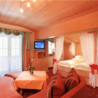 A cozy room with wooden walls and a double bed surrounded by curtains. In the foreground, there is a seating area with a table and a television.