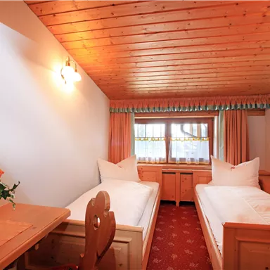 A cozy bedroom with two single beds and a wooden table. The room has wooden ceilings and large windows that provide natural light.