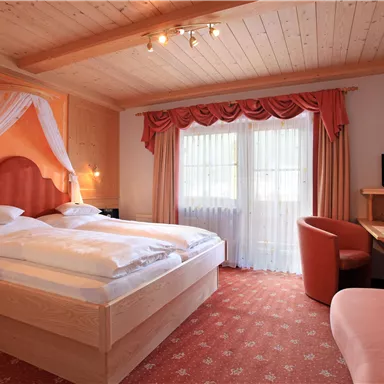 A cozy bedroom with a large double bed and light wooden furniture. Also visible are a table, a chair, and a window with curtains.