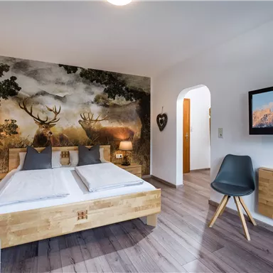 Ein modernes Schlafzimmer mit einem Holzbett und einer stilvollen Wandgestaltung mit Wildtieren. Der Raum ist hell und gemütlich mit einem Fernseher und einer Sitzgelegenheit.