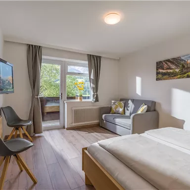 Ein helles Zimmer mit einem Doppelbett, einem Sofa und einem Fernseher. Große Fenster sorgen für viel Tageslicht und einen Blick nach draußen.