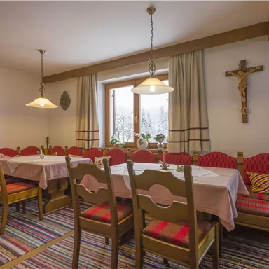 A cozy dining hall with long tables and red upholstered chairs. A cross hangs on the wall and large windows let in plenty of light.