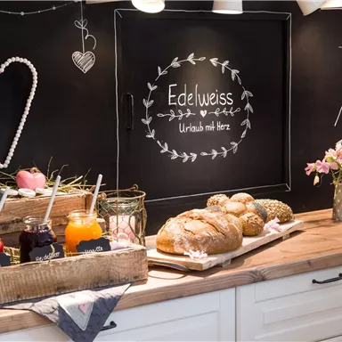 Eine gemütliche Küche mit frischen Brötchen und verschiedenen Marmeladen in Holzkisten. An der Wand steht die Aufschrift „Edelweiss“ und ein dekoratives Herz.