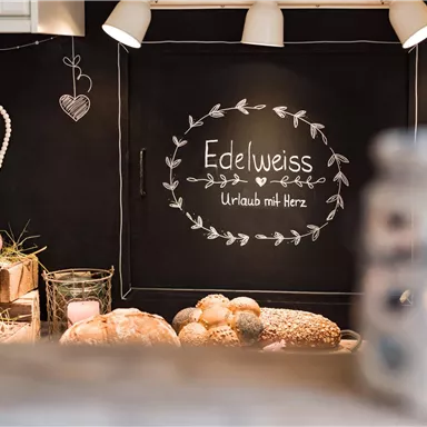 Eine einladende Küche mit einer Tafel, auf der "Edelweiss" und "Urlaub mit Herz" steht. Neben frischem Brot und Getränken ist die Atmosphäre warm und gemütlich.