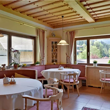 A cozy interior with round tables and a wooden ceiling. Large windows provide a view of the nature outside.