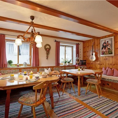 Ein gemütlicher Speisesaal mit Holzwänden und einem großen Tisch. Bunte Vorhänge und ein Teppich verleihen dem Raum eine warme Atmosphäre.