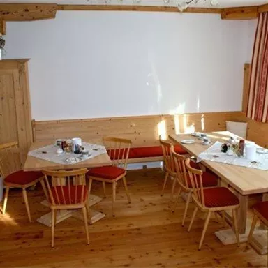 Ein gemütlicher Speiseraum mit Holztischen und Stühlen. Die Wände sind hell und es gibt Pflanzen im Raum.