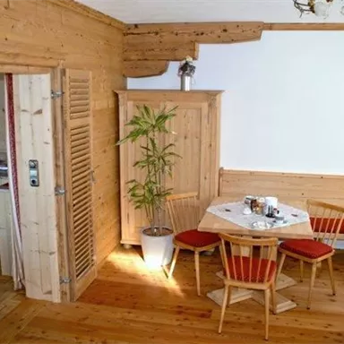 Eine gemütliche Stube mit Holzverkleidung und modernen Möbeln. Am Tisch stehen Stühle bereit, während eine Pflanze im Raum für eine grüne Note sorgt.