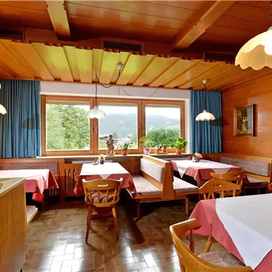 A cozy room with wooden furniture and colorful tablecloths. Large windows offer a view of the greenery and let in plenty of daylight.