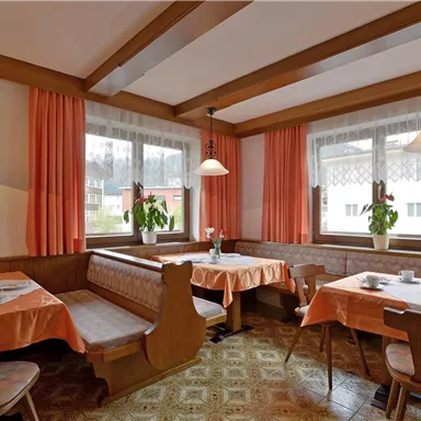 A cozy dining room with wooden furniture and orange tablecloths. The windows are decorated with curtains, and there are plants on the tables.