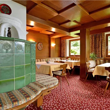 A cozy restaurant with light wooden interior and traditional tables. A beautiful tiled stove and windows that let in plenty of daylight.