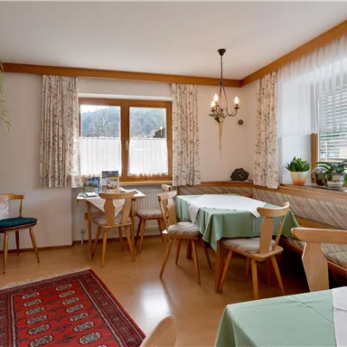 Ein helles Esszimmer mit Holzmöbeln und einem großen Fenster. Die Tische sind gedeckt und Pflanzen bringen eine grüne Note in den Raum.