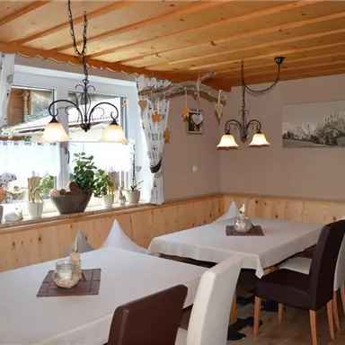 Ein gemütlicher Speisebereich mit Holzvertäfelung und großen Fenstern. Die Tische sind einfach gedeckt, und die Wände sind mit Bildern und Pflanzen dekoriert.