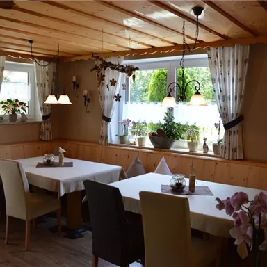 Ein gemütliches Esszimmer mit hellen Tischen und Stühlen. Die Fenster sind mit Vorhängen geschmückt, und Pflanzen sorgen für eine einladende Atmosphäre.