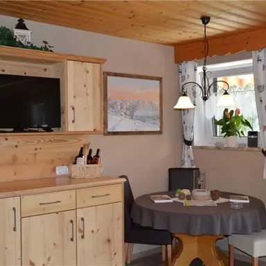 Eine gemütliche Küche mit Holzschränken und einem Tisch. Ein Fernseher hängt an der Wand und ein Fenster mit Vorhängen lässt natürliches Licht herein.