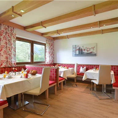 Ein gemütlicher Speisesaal mit roten Sitzbänken und Tischdecken. Die Fenster bieten einen Blick ins Grüne und der Raum ist freundlich dekoriert.