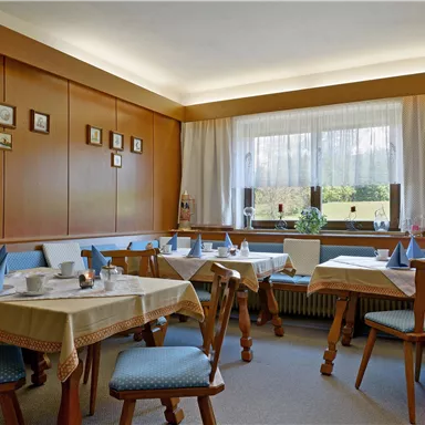 Ein gemütlicher Speiseraum mit hellen Tischen und Stühlen. Durch die Fenster fällt Licht auf die dekorierte Wand.