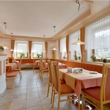 Ein gemütliches Restaurant mit vielen Tischen und Stühlen. Fenster mit Vorhängen und Pflanzen sorgen für eine einladende Atmosphäre.