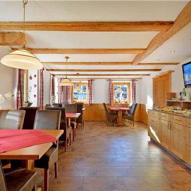 Ein gemütlicher Essbereich mit Holzmöbeln und roten Vorhängen. Auf dem Buffet sind diverse Speisen angerichtet, und ein Fernseher zeigt ein Programm.