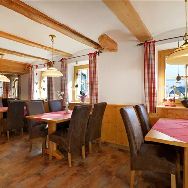 Eine gemütliche Gaststube mit Holztischen und Stühlen. Rote Tischdecken und traditionelle Fensterdekorationen sorgen für eine warme Atmosphäre.