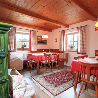 A cozy dining room with wooden beam ceiling and warm colors. There are reddish tablecloths and a nice stove in the corner.