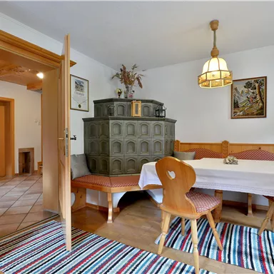 A cozy dining area with a wooden table and chairs. In the background, there is a traditional stove and the walls are paneled with wood.
