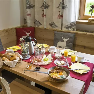 Ein schön gedeckter Frühstückstisch in einer gemütlichen Umgebung. Es gibt frische Brötchen, Getränke und ein reichhaltiges Frühstücksgericht.