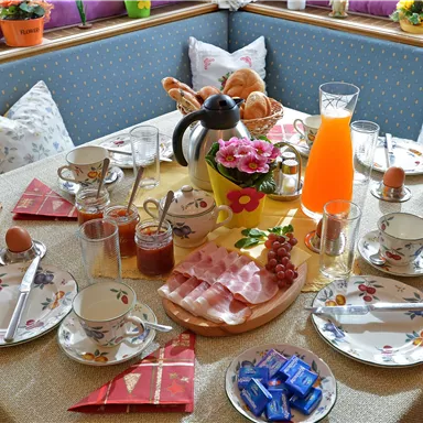 Ein gemütlicher Frühstückstisch mit verschiedenen Speisen. Darunter befinden sich Aufschnitt, Brötchen, Eier und Getränke in ansprechenden Tassen und Gläsern.