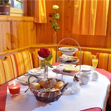A cozy table in a wooden room, decorated with a rose and fresh fruit. On the table are rolls, croissants, and a glass of juice arranged.