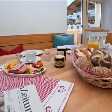 A cozy breakfast table with fresh rolls, sausage, cheese, and fruit. A bouquet of flowers and a newspaper complete the picture.