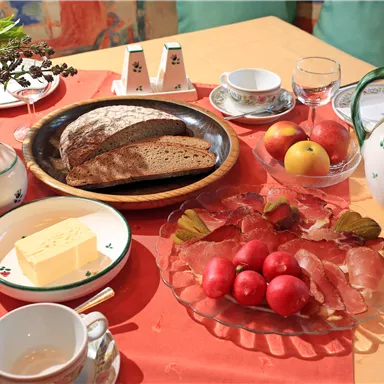 Ein Tisch gedeckt mit verschiedenen Köstlichkeiten. Darauf liegen Brot, Käse, Obst und Wurst auf einer ansprechenden Tischdecke.