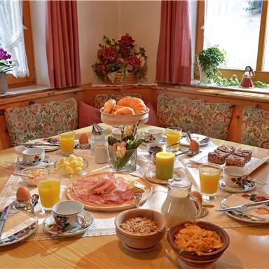 Ein gemütlicher Frühstückstisch mit verschiedenen Speisen, darunter Obst, Brötchen, Wurst, Käse und Getränke. Die Atmosphäre ist einladend mit Blumen und Tageslicht.