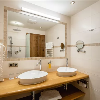 Ein modernes Badezimmer mit zwei Waschbecken und einem großen Spiegel. Die Wände sind in hellen Tönen gehalten und es gibt eine Dusche im Hintergrund.