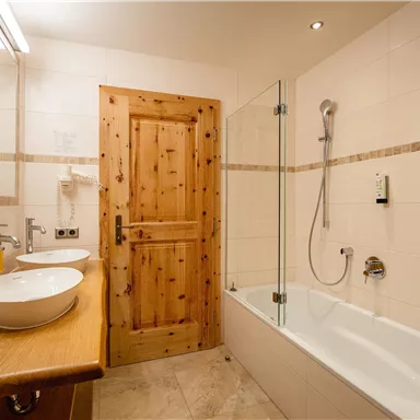 Ein modernes Badezimmer mit einer Badewanne und zwei Waschbecken. Die Wände sind hell gefliest, und eine Holzde Tür verleiht dem Raum eine warme Atmosphäre.