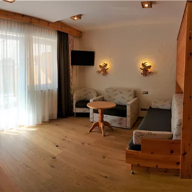 Ein gemütlicher Raum mit Holzverkleidung, einem Tisch und zwei Sesseln. Große Fenster lassen viel Tageslicht hereinkommen.