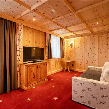 Ein gemütlicher Raum mit Holzwänden und einem roten Teppich. Er verfügt über ein Sofa, einen Fernseher und große Fenster, die Tageslicht hereinlassen.