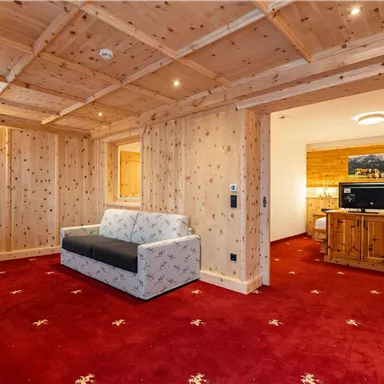 Ein gemütliches Zimmer mit Holzwänden und rotem Teppich. Es gibt ein Sofa, einen Fernseher und ein angrenzendes Schlafzimmer.