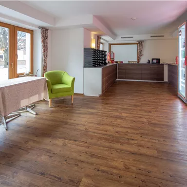 Ein modern gestalteter Empfangsbereich mit hellem Holzfußboden. Zwei grüne Stühle stehen vor großen Fenstern, und der Empfangstresen ist im Hintergrund sichtbar.