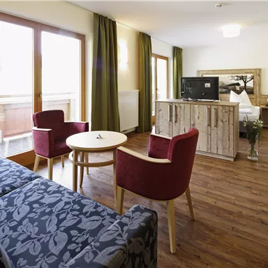 Ein gemütliches Zimmer mit einem Wohnzimmerbereich, einem Tisch und zwei Stühlen. Große Fenster lassen viel Licht herein und bieten einen Blick auf den Balkon.