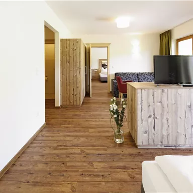 Ein modernes Zimmer mit Holzfußboden und einer gemütlichen Einrichtung. Der Raum verfügt über einen Fernseher und große Fenster mit grünen Vorhängen.