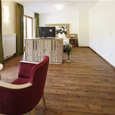 Ein modernes Zimmer mit Holzboden und großen Fenstern. Es gibt einen roten Stuhl, einen Tisch und einen Fernseher auf einer Holzkommode.