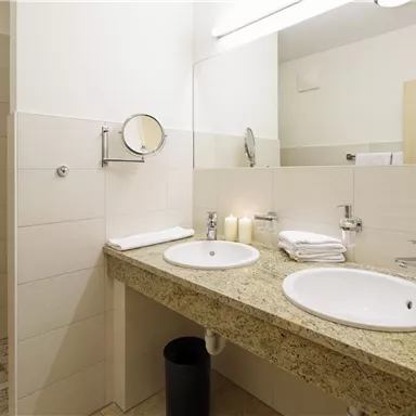 Ein modernes Badezimmer mit zwei Waschbecken und einem großen Spiegel. Die Wände sind hell gefliest, und ein Handtuchhalter ist sichtbar.