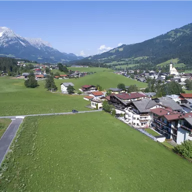 Eine malerische Landschaft mit grünen Wiesen und kleinen Häusern. Im Hintergrund sind Berge und ein klarer Himmel zu sehen.