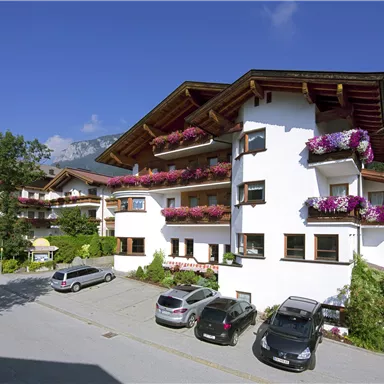 Ein gepflegtes Gebäude im Alpenstil mit bunten Blumenbalkonen. Umgeben von Bäumen und einer ruhigen Straße mit parkenden Autos.