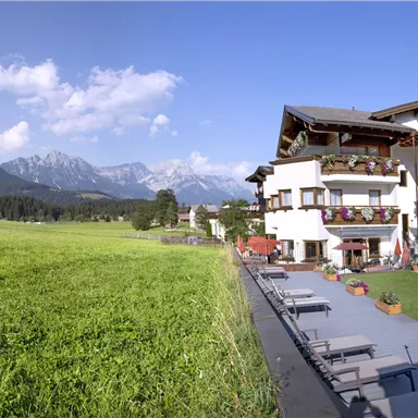 Ein malerisches Hotel mit Balkonen, umgeben von grünen Wiesen und Bergen im Hintergrund. Der Himmel ist klar und blau, ideal für einen entspannten Aufenthalt.
