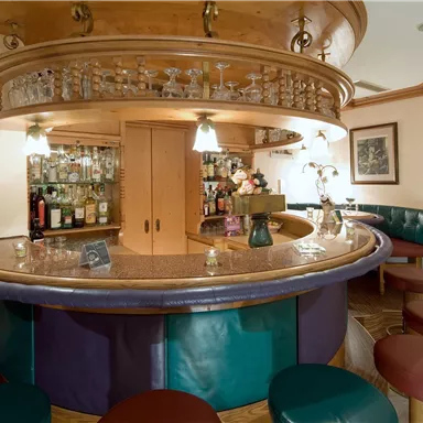 Eine gemütliche Bar mit einem runden Tresen und verschiedenen Sitzgelegenheiten. Im Hintergrund sind Regale mit Getränken und eine gedämpfte Beleuchtung zu sehen.