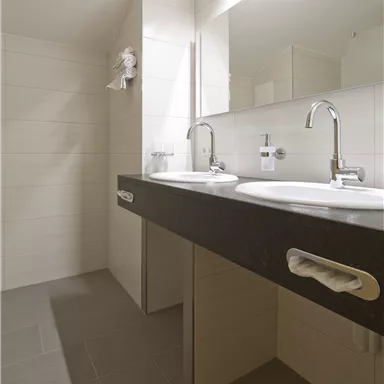 Ein modernes Badezimmer mit zwei Waschbecken und einem großen Spiegel. Die Wände sind hell und die Fliesen sind grau.